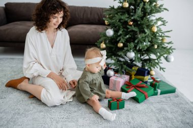 Neşeli anne, küçük kızın yanında oturmuş Noel ağacının yanındaki hediye kutusuna bakıyor.