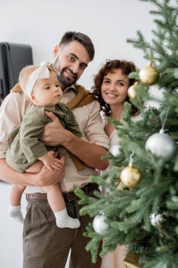 Mutlu bir baba, mutlu bir eş ve süslü bir Noel ağacının yanında küçük kızını tutuyor. 