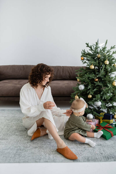 веселая мама сидит рядом с украшенной елкой с подарками и младенческой дочерью 