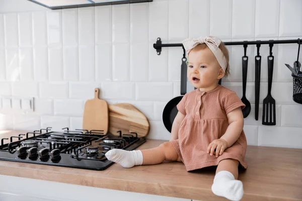 穿着发带和粉色衣服坐在炉边厨房工作台上的女婴感到惊讶 — 图库照片