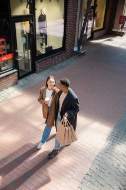 high angle view of interracial couple in trendy coats walking with shopping bags and coffee to go along showcases clipart