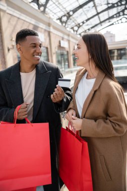 Neşeli Afro-Amerikalı adam, arka planda alışveriş merkezinin yakınında alışveriş torbaları tutan hayran kalmış kız arkadaşına kredi kartı gösteriyor.