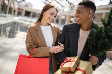 Küçük bir Noel ağacı ve hediye kutusuyla şık bir afro-amerikan erkeği alışveriş çantalı kız arkadaşına bakıyor.