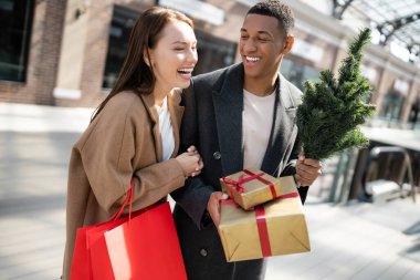 Alışveriş torbalarıyla heyecanlı bir kadın şık bir Afrikalı Amerikalının yanında gülüyor. Elinde hediyeler ve küçük bir noel ağacı var.