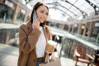 Alışveriş torbaları ve paket servis içkileriyle modaya uyan bir kadın dışarıda akıllı telefondan konuşuyor.