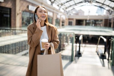 Alışveriş poşetleri ve kahveyle bulanık alışveriş merkezinin yakınına giderken cep telefonuyla konuşan heyecanlı bir kadın.