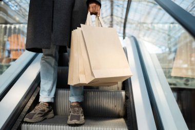 Kürklü ve spor ayakkabılı Afro-Amerikan bir adamın yürüyen merdivende alışveriş torbası tutuşu.