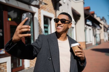 Güneş gözlüklü, şık ve neşeli Afro-Amerikan adam elinde kağıt bardak ve bulanık sokakta akıllı telefondan selfie çekiyor.