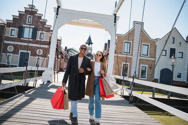 Cheerful Multiethnic Couple Stylish Clothes Sunglasses Walking Shopping Bags Bridge — Stock Photo, Image