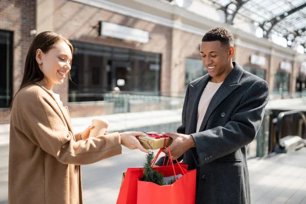 快乐的女人拿着纸杯从时髦的非洲男人手里接过礼品盒 拿着购物袋 — 图库照片