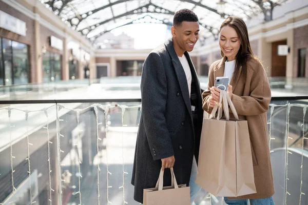 Smiling Interracial Couple Trendy Coats Holding Shopping Bags Looking Smartphone — Stock Photo, Image