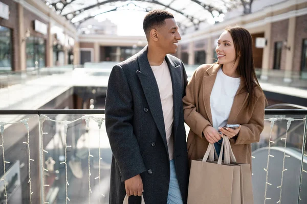 一个漂亮的女人 拿着手机和购物袋 在模糊的购物中心附近对着时髦的非洲裔美国男人微笑 — 图库照片