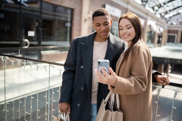 朗らかなブルネットの女性ショッピングバッグを持つ流行のアフリカ系アメリカ人男性の近くのスマートフォンを見て — ストック写真