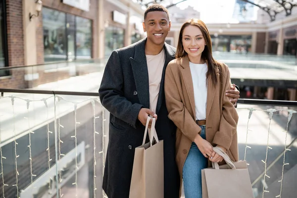 Cheerful Multiethnic Couple Trendy Coats Holding Shopping Bags Looking Camera — Stock Photo, Image