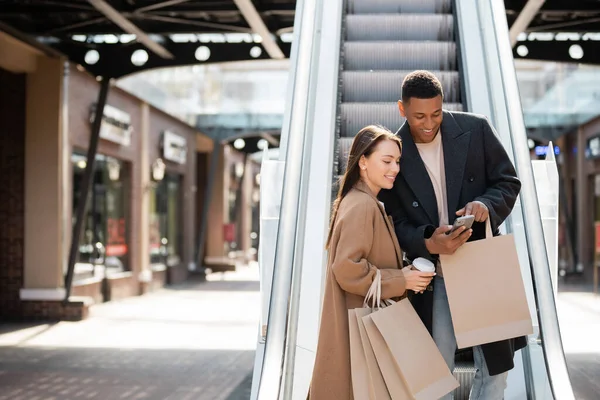 美国黑人男子指着智能手机 微笑着 拿着购物袋和纸杯站在自动扶梯上 — 图库照片