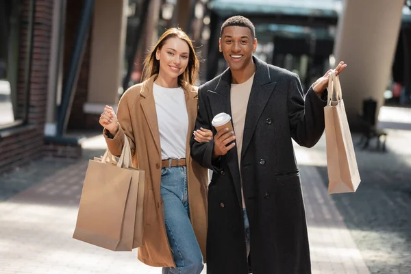 stylish multiethnic couple with shopping bags and coffee to go smiling at camera on blurred street