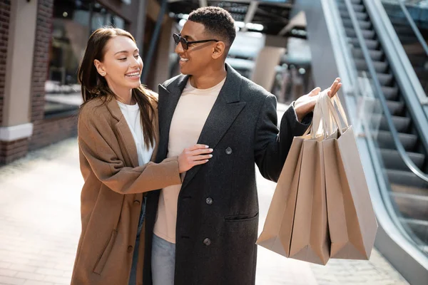 african american man in sunglasses holding shopping bags near happy girlfriend on street