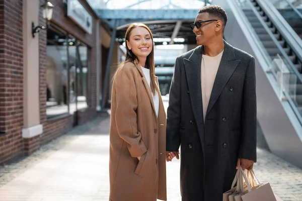 pleased woman with hand in pocket of coat holding hands with stylish african american man with shopping bags