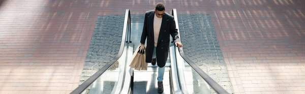высокий угол зрения на модный африканский американец с сумками для покупок на эскалаторе, баннер
