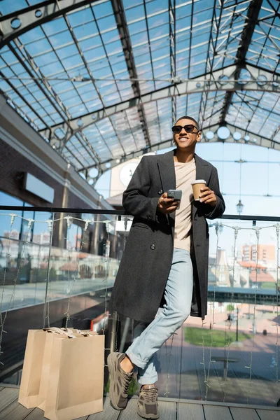 커피를 아프리카 미국인 남자와 쇼핑백 쇼핑몰 근처에 스마트폰 — 스톡 사진