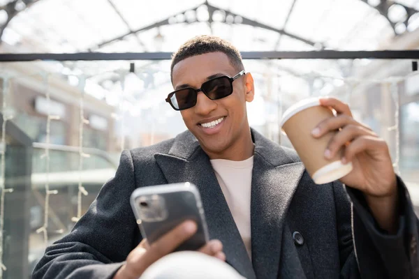 幸せなアフリカ系アメリカ人男性サングラス保持テイクアウトドリンクや携帯電話を見て屋外 — ストック写真