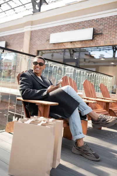 African American Man Coat Sunglasses Sitting Smartphone Shopping Bags Mall — Stock Photo, Image