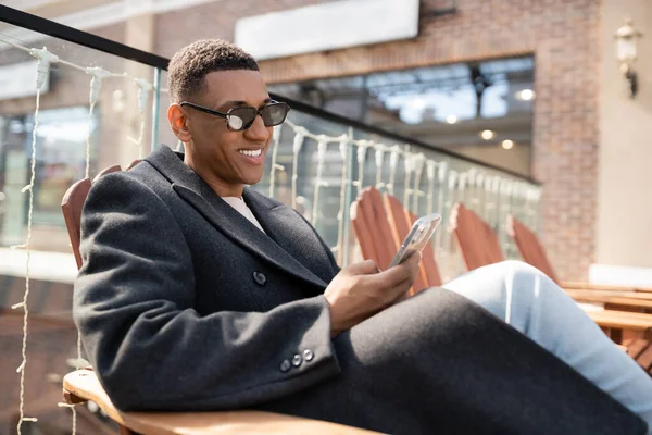 Glimlachende Afro Amerikaanse Man Trendy Jas Zonnebril Chatten Mobiele Telefoon — Stockfoto