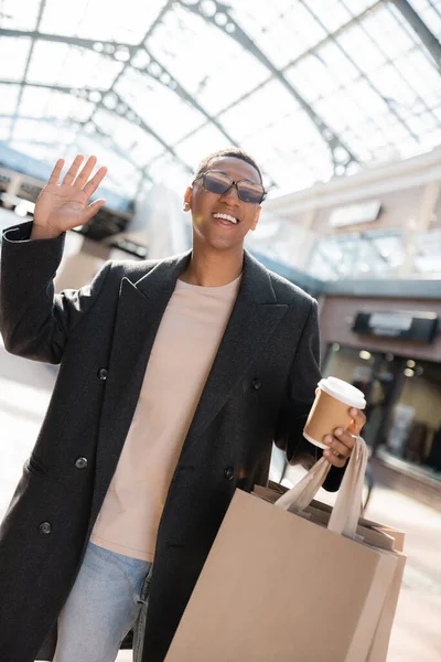 joyful african american man with paper cup and shopping bags waving hand on city street