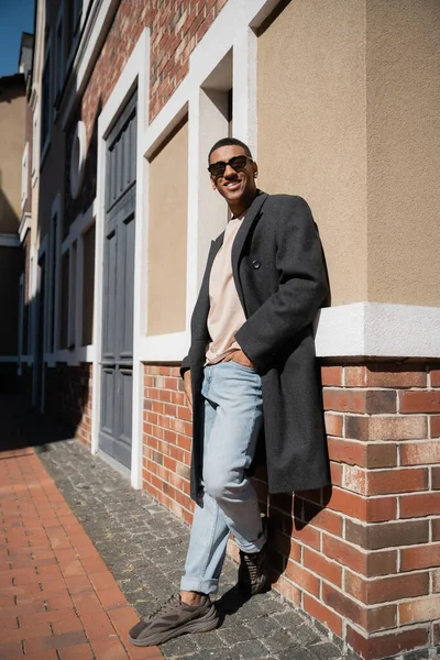 コートを着たアフリカ系アメリカ人男性と建物の近くのジーンズのポケットに手を入れて立つサングラスの長さ — ストック写真