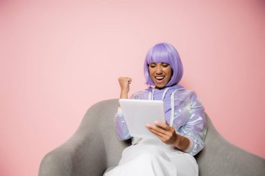 Heyecanlı, mor peruklu, dijital tablet kullanan ve pembe koltukta tek başına oturan Afro-Amerikan kadını. 