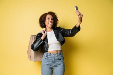 Şık deri ceketli mutlu Afro-Amerikalı kadın alışveriş torbaları tutuyor ve sarıya selfie çekiyor.