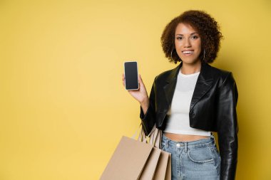 Deri ceketli mutlu Afro-Amerikalı kadın elinde alışveriş çantaları ve üzerinde sarı ekran olan cep telefonu var.