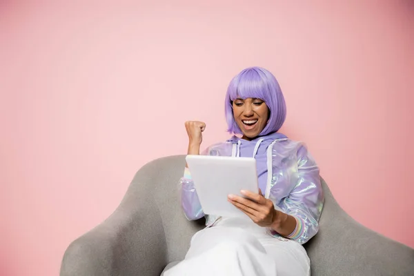 紫のウィッグを着たアフリカ系アメリカ人の興奮した女性がデジタルタブレットを使ってピンクの椅子に座り — ストック写真
