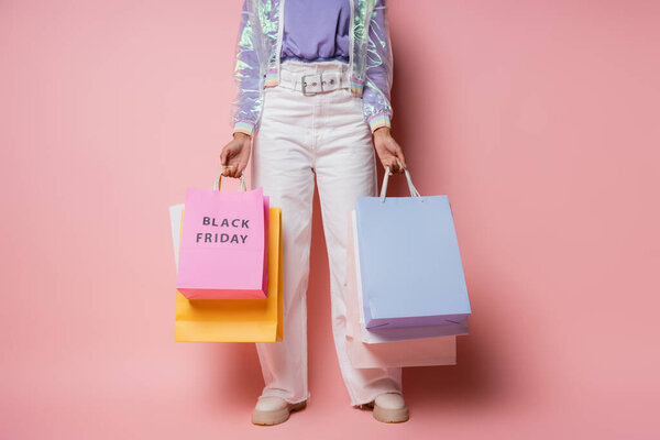 cropped view of woman in transparent jacket and white pants holding shopping bags with black friday lettering on pink 
