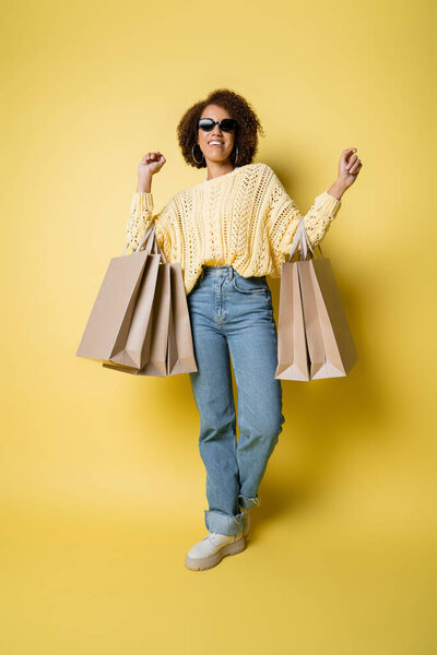 full length of cheerful african american woman in sunglasses holding shopping bags on yellow