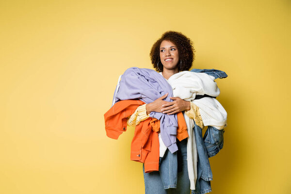 happy african american woman holding pile of colorful clothes on yellow background
