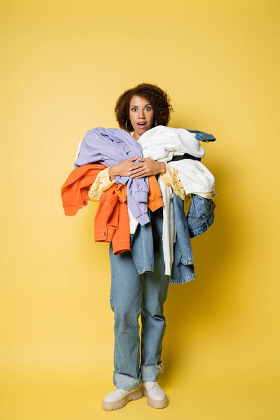 full length of shocked african american woman holding pile of colorful clothes on yellow background