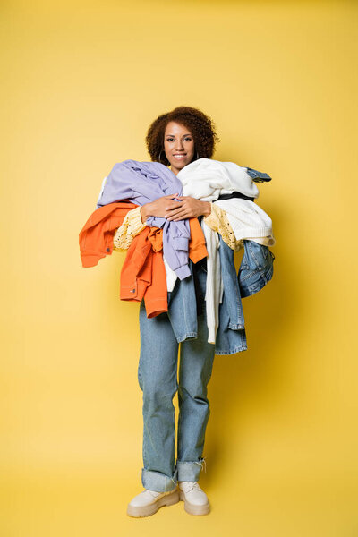 full length of happy african american woman holding pile of colorful clothes on yellow 