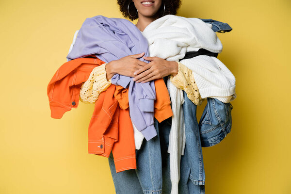cropped view of happy african american woman holding pile of colorful clothes on yellow 
