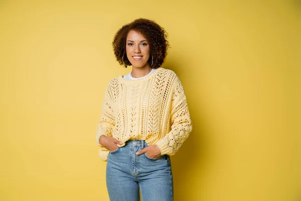 Szczęśliwa Młoda Afroamerykanka Dzianinowym Swetrze Stojąca Rękami Kieszeniach Dżinsach Żółtych — Zdjęcie stockowe