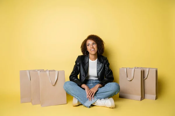 黄色のショッピングバッグの近くに座っている革のジャケットやジーンズで幸せなアフリカ系アメリカ人女性の完全な長さ — ストック写真