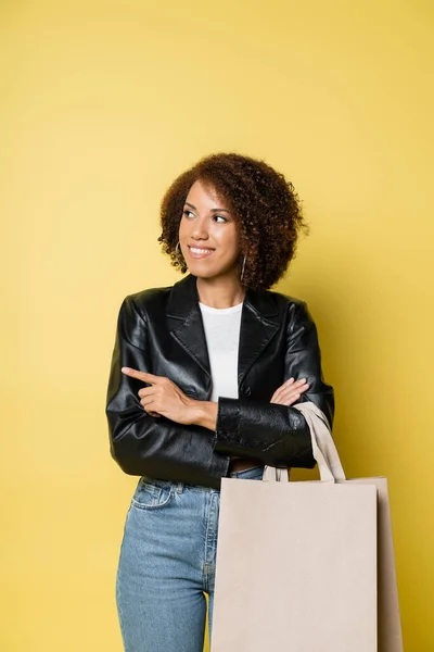 穿着时髦皮夹克的快乐的非洲裔美国女人拿着购物袋购物 用手指着黄色 — 图库照片