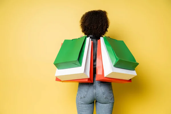 back view of curly african american woman in stylish blue jeans holding black friday shopping bags on yellow