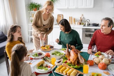 Çok ırklı ailelerin yanında pasta kesen gülümseyen bir kadın ve evde yemek yerken çocuklar. 