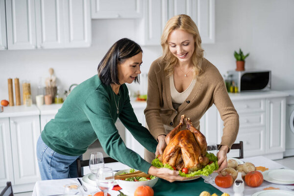 Positive interracial mother and adult daughter putting thanksgiving turkey on table 