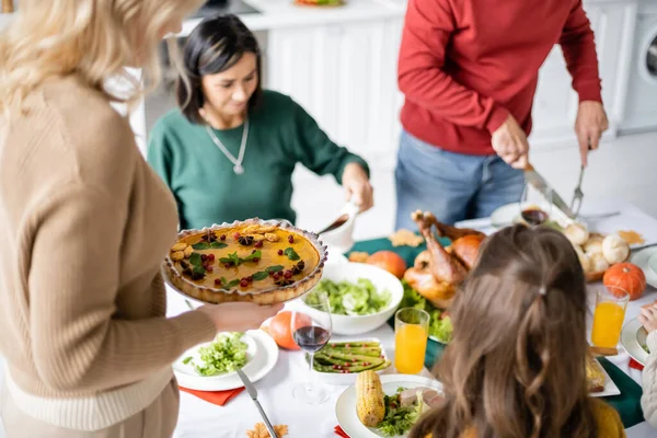 Woman holding pie near blurred family and thanksgiving dinner at home