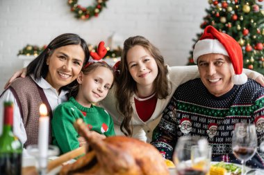 Irklar arası büyükanneler ve torunlar, bulanık ön planda, enfes bir Noel yemeğinin yanında kameraya gülümsüyor.