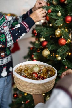 Hasır sepet taşıyan bir kadının, kocasının yanında Noel ağacı süsleyen mücevherlerle dolu bir manzarası.