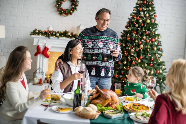 smiling man toasting with glass of red wine during festive dinner with interracial family near decorated christmas tree