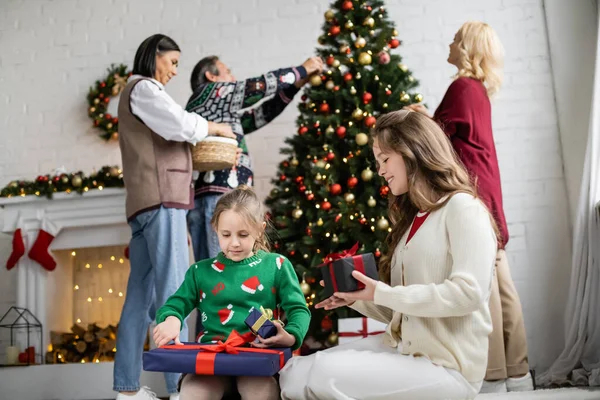 girls looking at new year presents while interracial family decorating christmas tree on blurred background